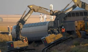 Workers stand on a train car on its side as front-loaders prop up another train car