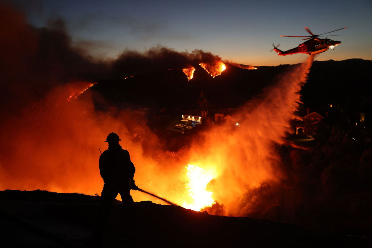 <i>David Swanson/AFP/Getty Images via CNN Newsource</i><br/>Fire personnel respond to homes destroyed while a helicopter drops water as the Palisades Fire burns on January 7.