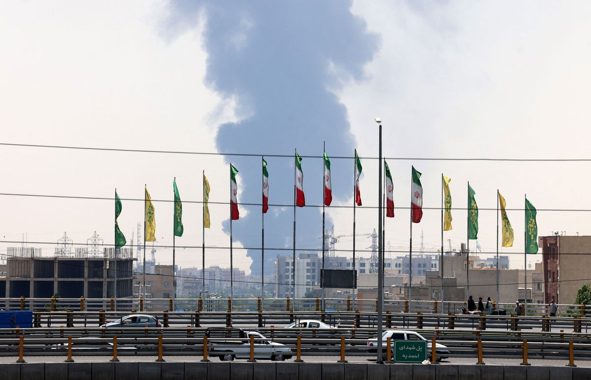 <i>Atta Kenare/AFP via Getty Images via CNN Newsource</i><br/>Flags flutter along a bridge as a plume of heavy smoke and fire rises from an oil refinery in southern Tehran on June 15